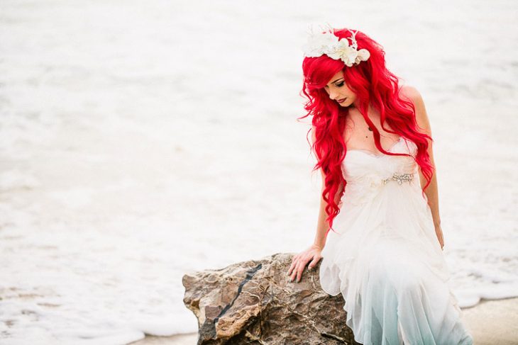 mujer con vestido blanco y azul y de cabello rojo sentada en una gran roca y tras ella se encuentra el mar 