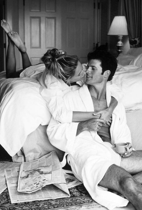 hombre y mujer, ella recostada en la cama y el sentado en el suelo mientras se abrazan y toman café