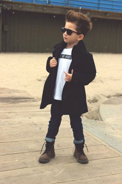 Niño usando una gabardina, botas y pantalón de mezclilla posando para una foto 