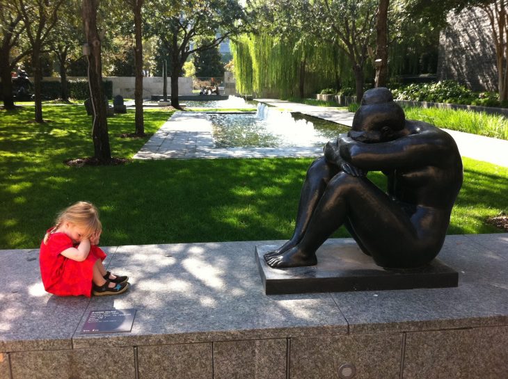 Niña sentada frente a una estatua con las manos en la cabeza