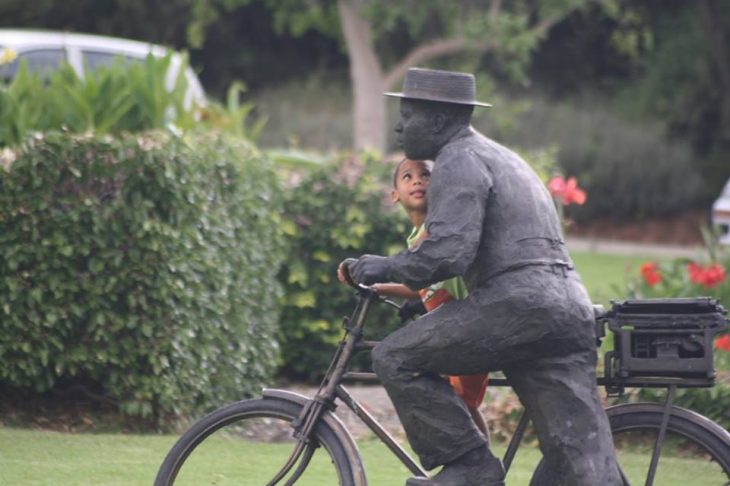 Niño sujetándose de la bici de un señor que es una estatua 