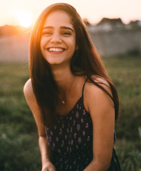 Chica de cabello castaño, lacio y largo sonriendo en el campo