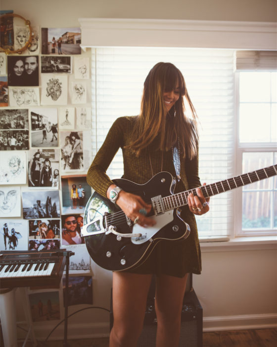 Chica de cabello castaño, lacio, largo con fleco, sonriendo, tocando la guitarra en su cuarto, pared llena de fotografías y dibujos
