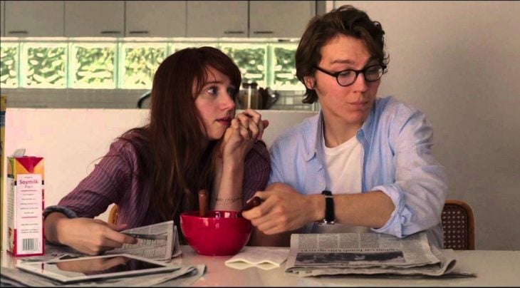 pareja de novios sentados en la mesa mientras el come cereal y lee el periódico y ella le toma la mano 