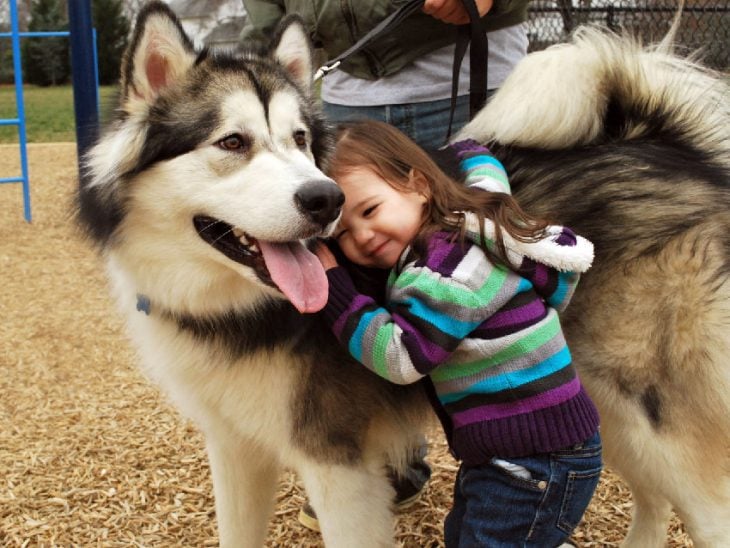 Una niña de pie recargada a un costado de su perro 