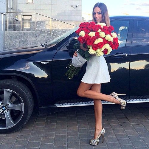 chica vestida de blanco sosteniendo un ramo de rosas frente a una camioneta negra 