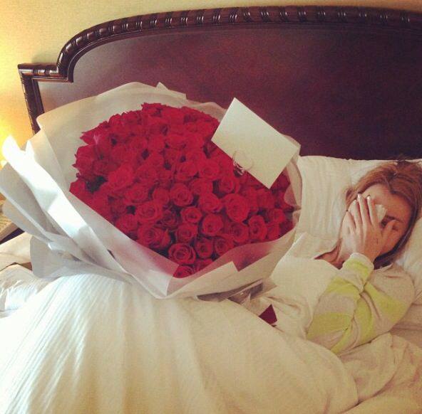 chica recostada en la cama mientras se sorprende al recibir rosas rojas con una tarjeta 