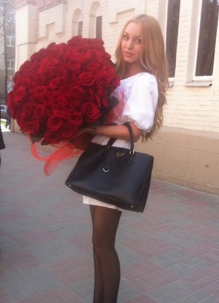 mujer en medio de la calle parada sosteniendo un gran ramo de rosas y su bolsa a un lado de ella 