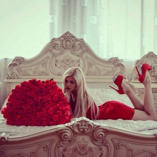 mujer vestida de rosjo acostada en medio de una cama blanca y frente a ella un ramo de rosas rojas 