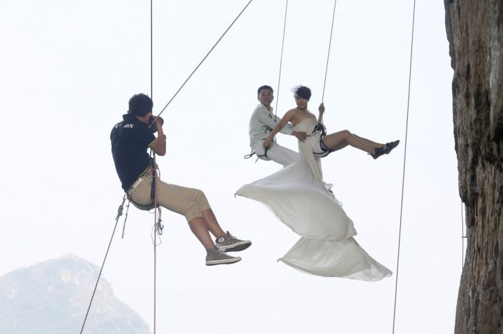 novios siendo fotografiados mientras cuelgan en un acantilado