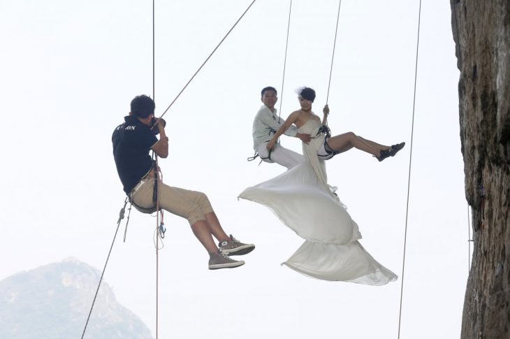 novios siendo fotografiados mientras cuelgan en un acantilado 