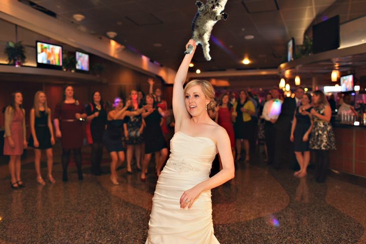 Novia en medio de una pista de baile mientras sostiene a un gato de una pata esperando lanzarlo a sus amigas que están a un lado de la barra de bebidas 