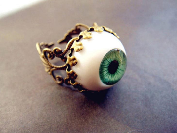 fotografía de anillo con un globo ocular en color blanco y verde y lineas doradas sobre una superficie de color café