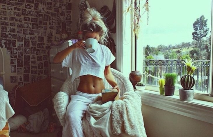 Chica enseñando el abdomen mientras toma café junto a la ventana de su casa