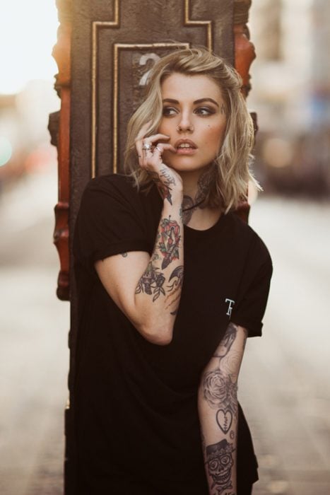 Chica con playera negra y tatuajes en los brazos mordiéndose un dedo
