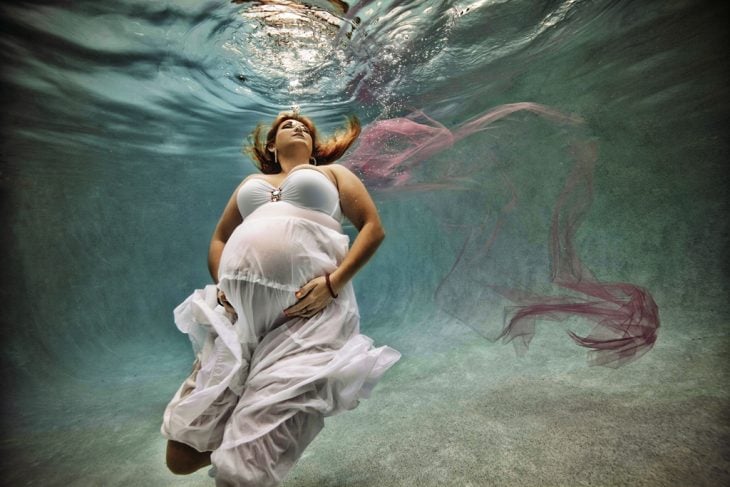 embarazada con vestido blanco tocándose el vientre bajo el agua 