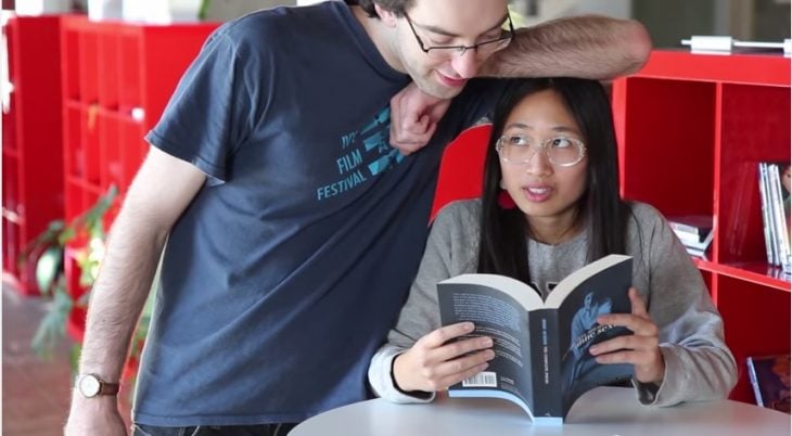mujer sentada leyendo un libro y hombre recargando su brazo en la cabeza de ella 