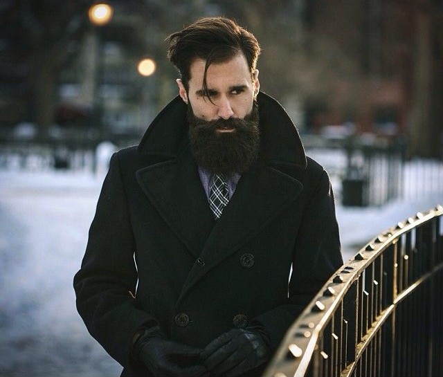 hombre con barba usando un saco parado a un lado de un puente 