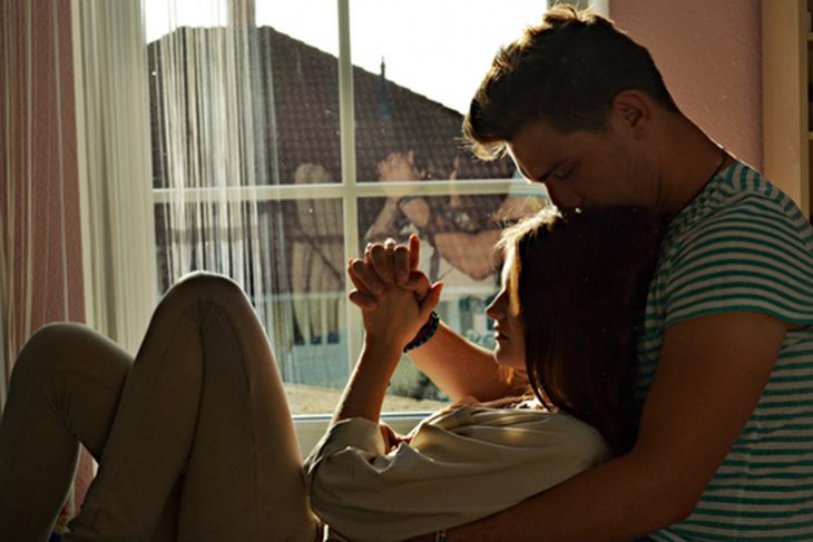 Chico abrazando a su novia al lado de una ventana 