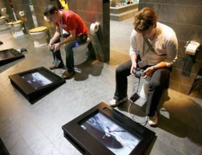 hombres sentados en la taza del baño y en el suelo una pantalla con controles para jugar vídeo juegos 