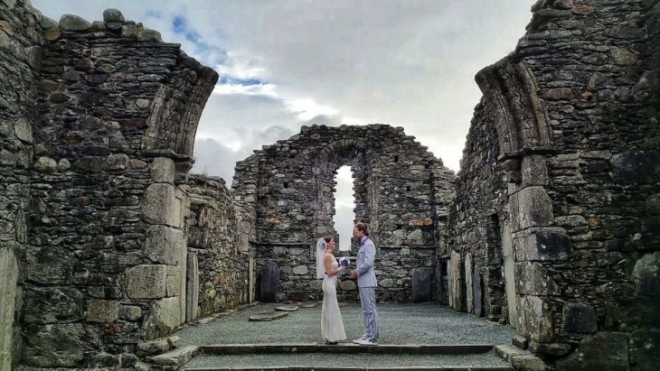 pareja parada frente a las ruinas de una antigua catedral en irlanda, vestidos para la ceremonia de su boda 