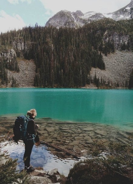 mujer parada a un lado de un lago con una mochila de viaje en sus hombros 