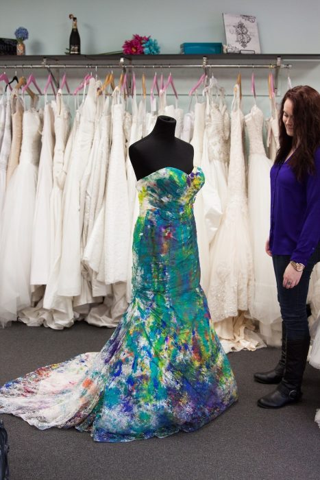 novia parada junto a su vestido de novia lleno de pintura que esta en una tienda de vestidos 