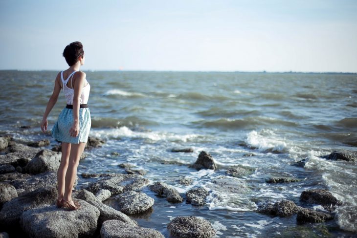 chica parada sobre unas rocas mirando hacia el mar 