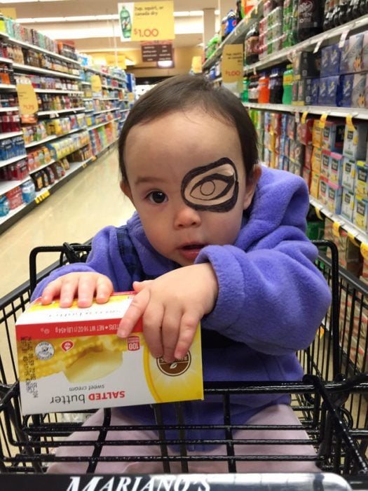 niña sentada en un carrito de super mercado usando un parche en el ojo 