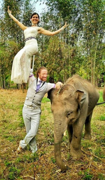 novio cargando a la novia en una mano y posando parados junto a un elefante 