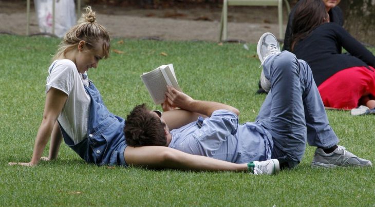 pareja de novios recostados en el pasto mientras él le lee un libro a ella 