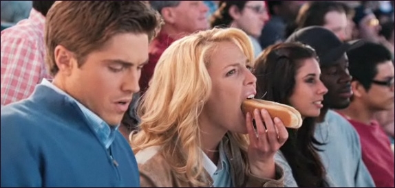 Escena de la película la cruda verdad protagonistas en un juego de beisbol comiendo 