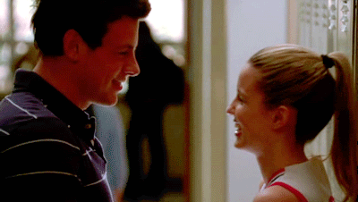 Gif glee chica y chico hablando mientras ela está recargada en la pared 