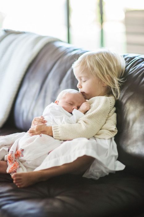 Niña besando en la frente a un bebé mientras ambos están senados en un sofá 