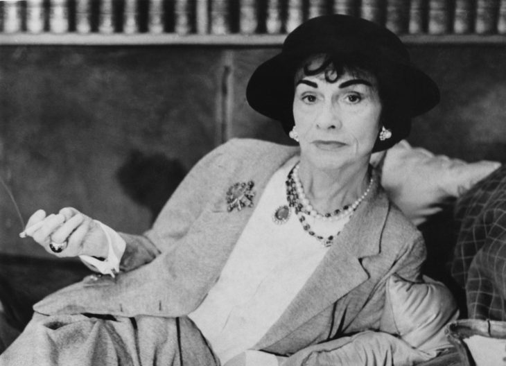 Diseñadora de modas Coco Chanel recostada sobre una cama fumando vistiendo un traje sastre y un sombrero 