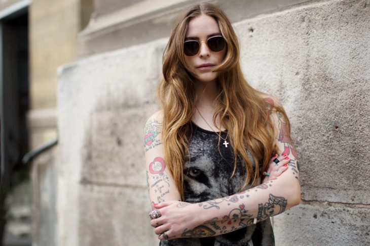 chica tatuada con lentes obscuros y cara seria recargada en la pared 