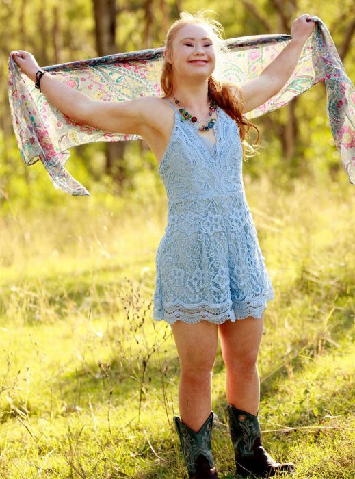 Niña con síndrome de down usando un vestido azul y botas alzando una mascada con sus manos 