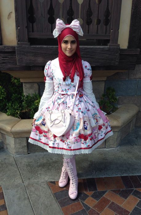 Chica usando un vestido de color blanco con flores rojas y un hijab en la cabeza y una bolsa blanca cruzada sobre el pecho 