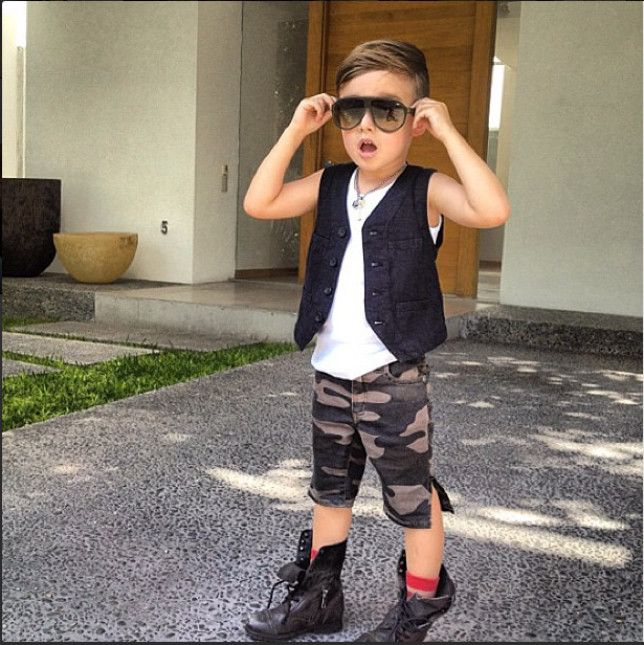 Niño usando un pantalón de militar corto, botas y chaleco parado a fuera de una casa 