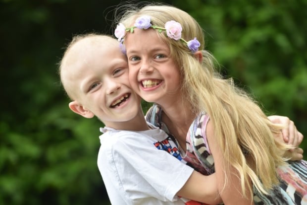 NIño con cáncer abrazado a una niña que tiene el cabello largo y rubio con una corona de flores 