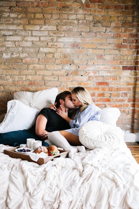 Pareja de novios recostados en la cama desayunando y compartiendo un beso 