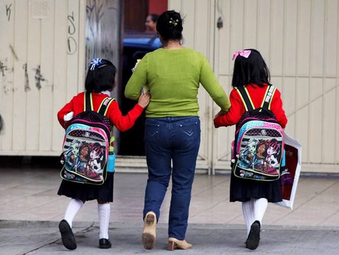 Niñas caminando de la mano de su mamá mientras van cargando mochilas para la escuela