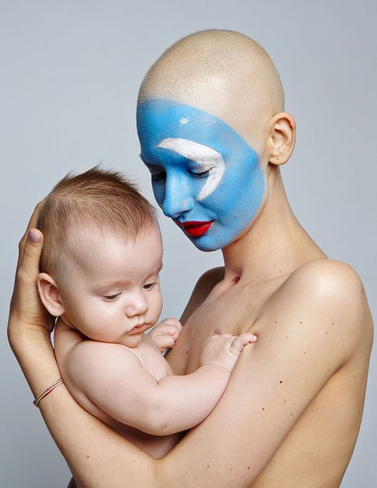 Modelo posando junto a su bebe mientras ella lo sostiene en brazos y con la cara pintada de azul 