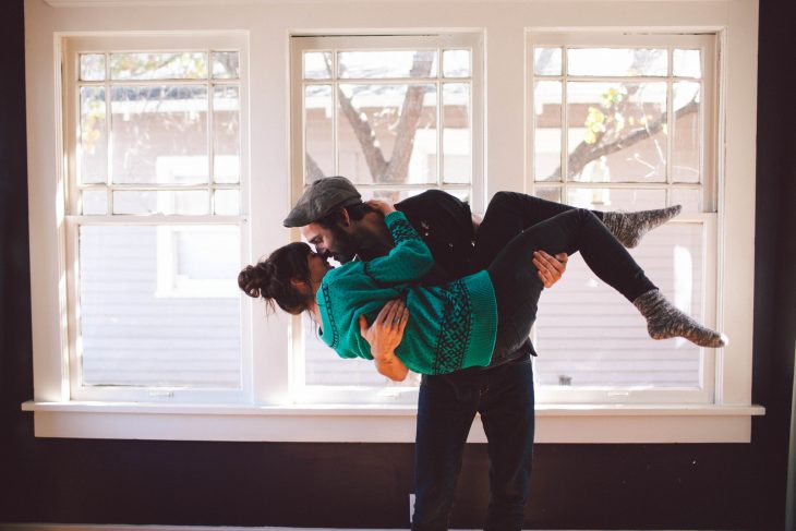 Hombre cargando a una chica para besarla mientras están en la sala de una casa