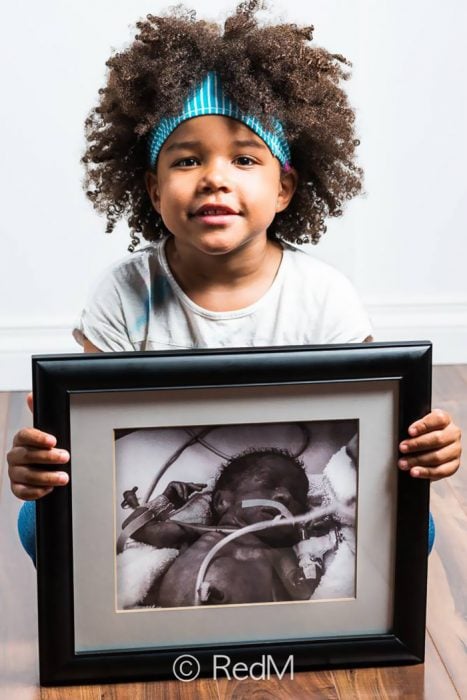 Niña sentada en el suelo sosteniendo una fotografía de ella cuando era bebé y estaba conectada a distintos tubos
