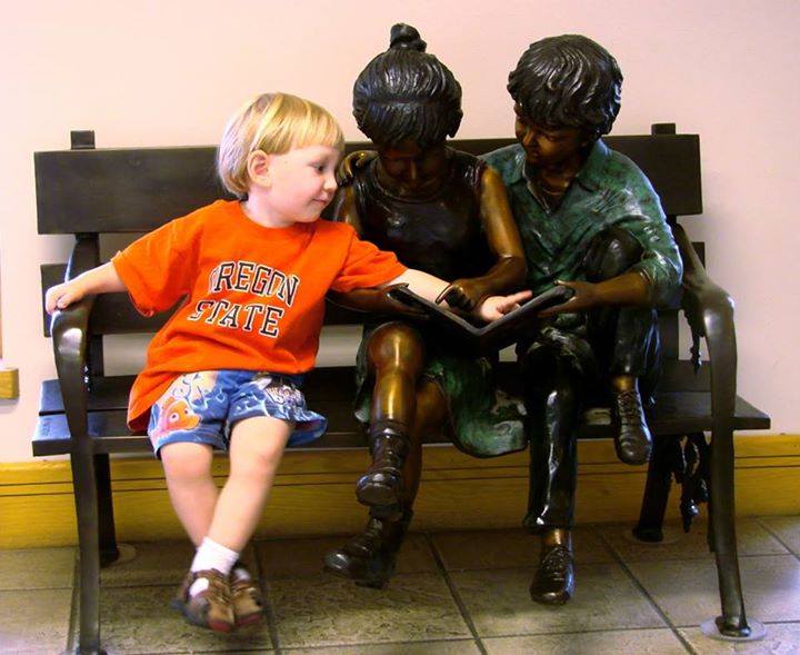 Niño intentando mostrar algo en un libro a unas estatuas