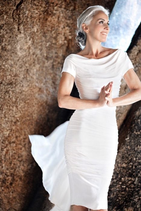 Mujer modelo de 59 años de edad usando un vestido de color blanco mientras está parada sobre las rocas