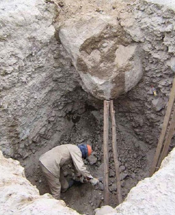 Hombre de bajo de una piedra que esta siendo sostenida con dos palos 