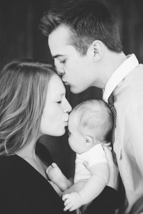 Hombre besando en la frente a una mujer mientras ambos cargan a una bebé