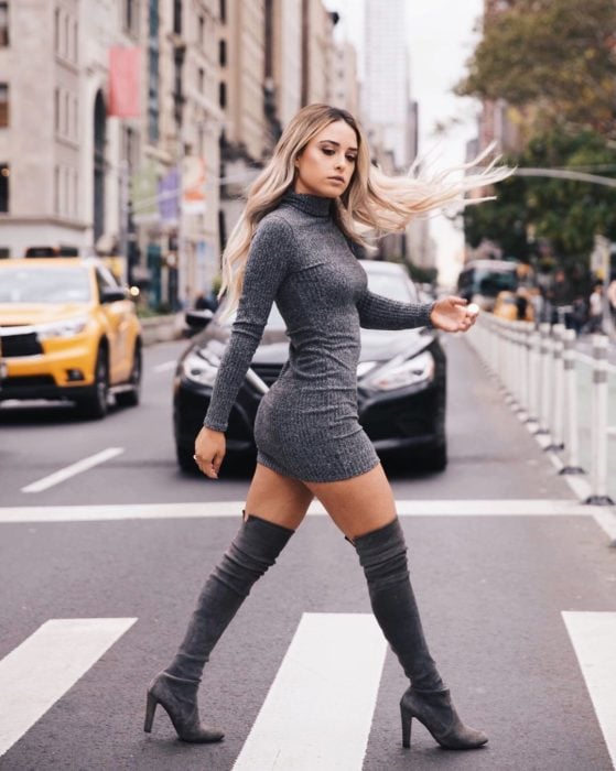Chica usando un vestido de color gris largo con botas altas mientras va caminando por la calle 
